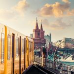 Ein gelber Zug fährt auf einer Hochbahn durch eine Stadt mit historischen Gebäuden unter einem bewölkten Himmel bei Sonnenuntergang.