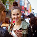 Zwei Frauen genießen aufgespießtes Essen auf einer belebten Straße mit bunten Schildern im Hintergrund.