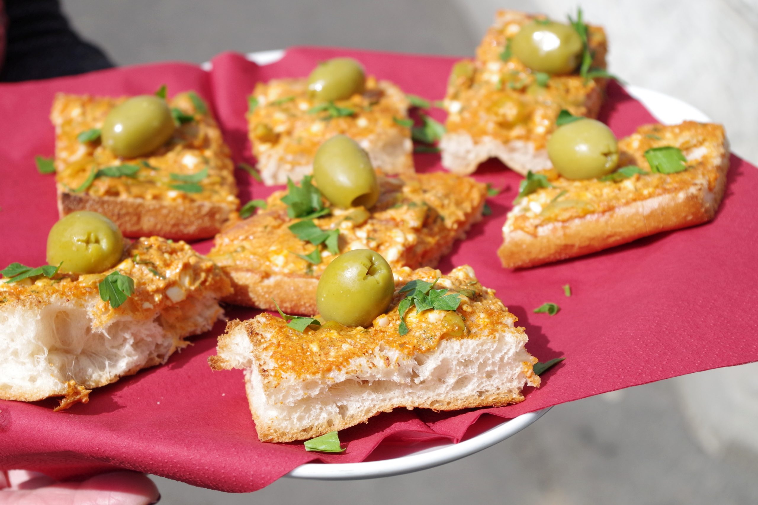 Mit Streichkäse, Oliven und Petersilie belegte Brotscheiben auf einem mit einer roten Serviette ausgelegten Teller, perfekt für eine Snackplatte zum Junggesellenabschied.