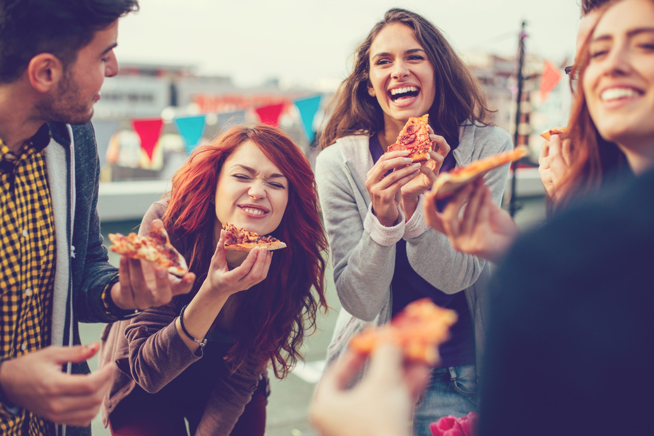 Eine Gruppe von Freunden lacht und genießt Pizza bei einem Treffen im Freien. Sie feiern einen Junggesellenabschied, während den Hintergrund festliche Wimpel schmücken.