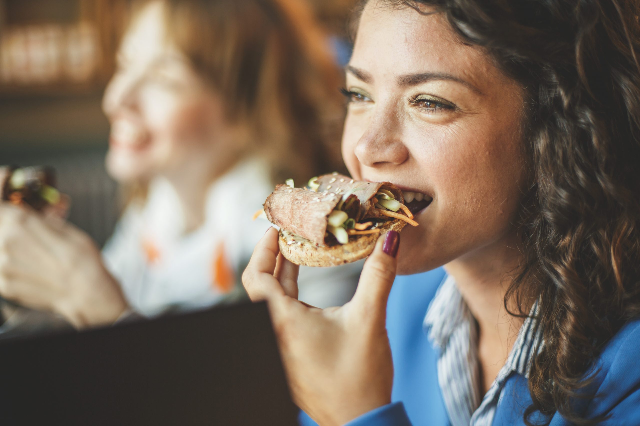 Eine Frau lächelt und isst ein Sandwich, im Hintergrund ist eine weitere Person verschwommen zu sehen. Die lebhafte Szene lässt an eine Junggesellenabschiedsfeier denken.