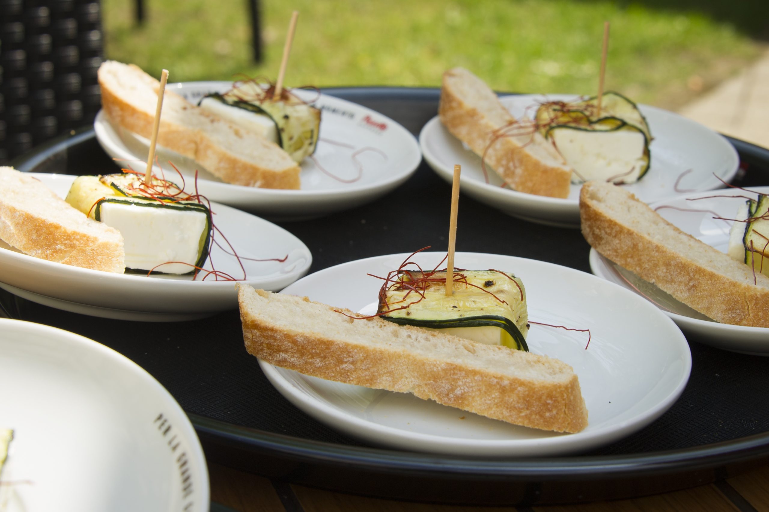 Bei einem malerischen Junggesellenabschied werden Teller mit Brotscheiben, Käse und mit dünnen Streifen garnierten Zucchini elegant im Freien auf einem Tablett serviert.