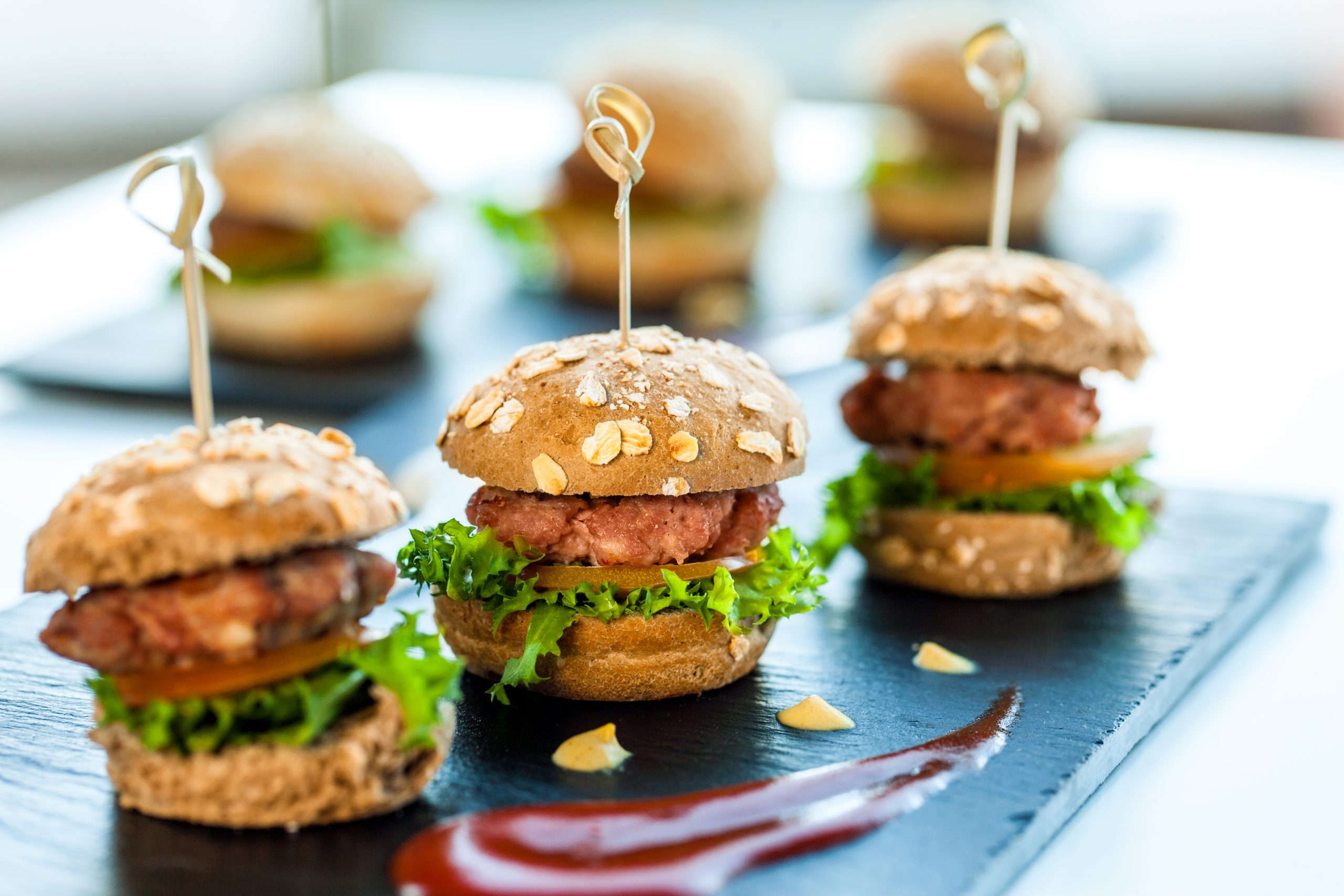 Mini-Burger mit Salat, Tomaten und Spießen zieren eine Schieferplatte, garniert mit Saucenwirbeln – ein perfekter Leckerbissen für jede Junggesellenabschiedsfeier.