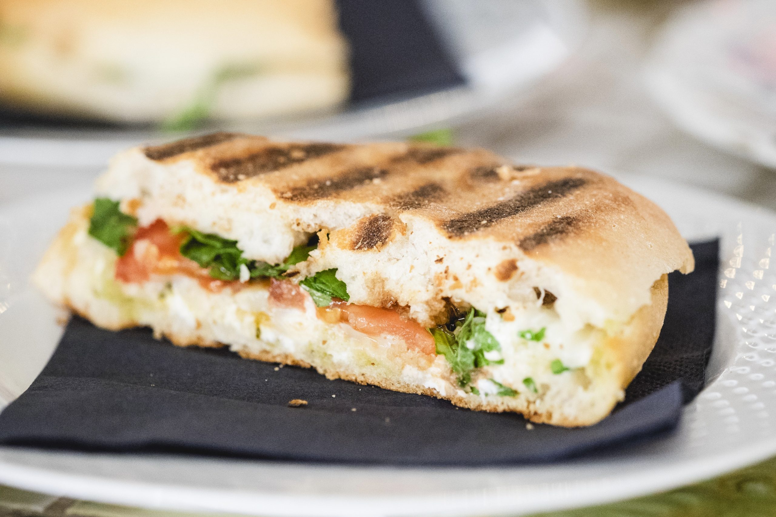 Ein gegrilltes Sandwich mit Tomaten, Gemüse und Käse liegt einladend auf einem weißen Teller auf einer schwarzen Serviette – perfekt, um vor den Abenteuern eines lebhaften Junggesellenabschieds Energie zu tanken.