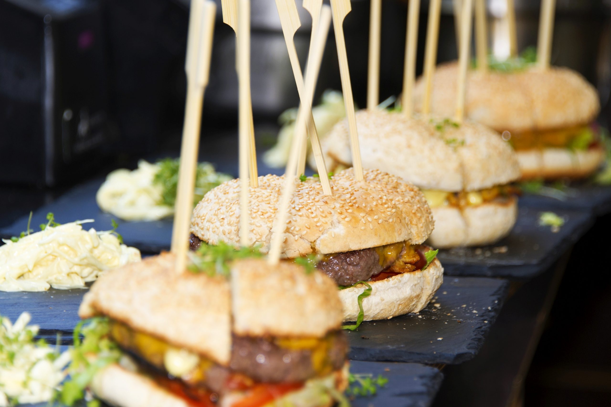 Vier Cheeseburger am Spieß auf schwarzen Tellern, garniert mit Gemüse und begleitet von Krautsalat, perfekt für ein Junggesellenabschied-Fest.