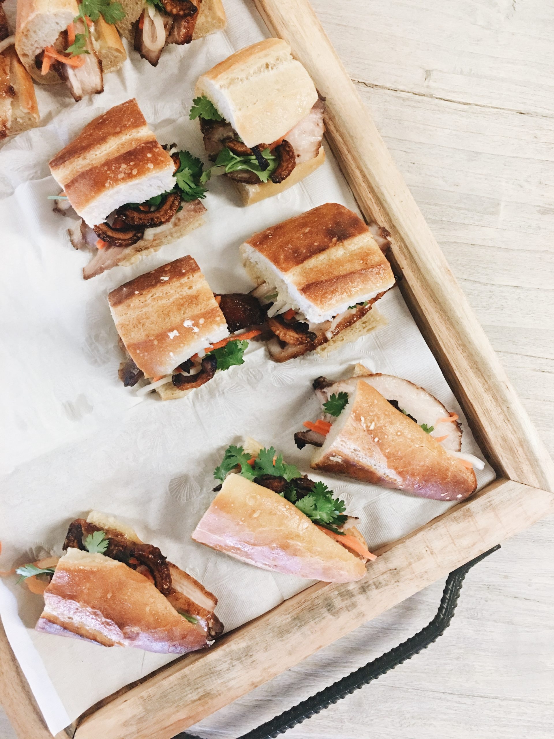 Ein Tablett mit Banh-Mi-Sandwiches mit Koriander steht auf einer hellen Holzoberfläche und ist perfekt, um einer lebhaften Junggesellenabschiedsfeier Energie zu verleihen.