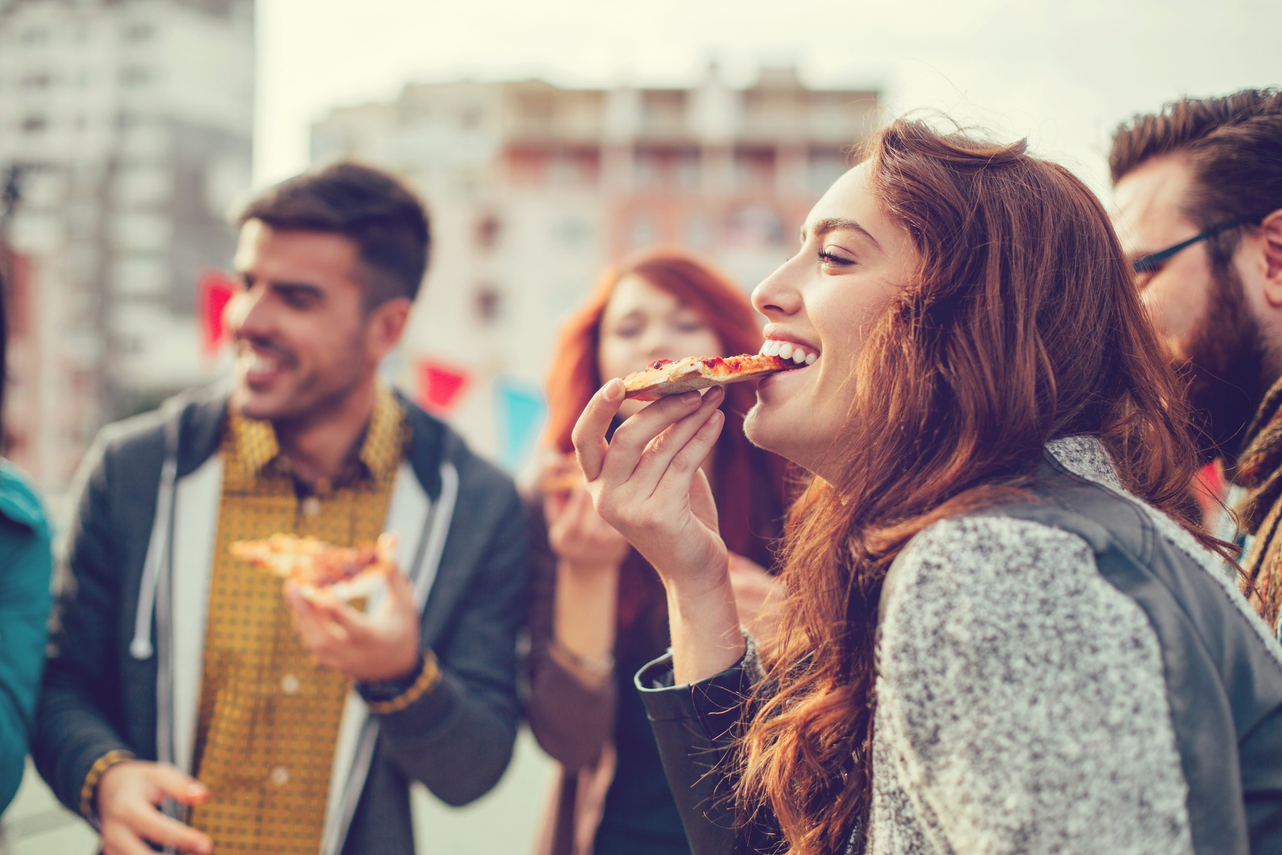 Eine Gruppe von Freunden, die einen lebhaften Junggesellenabschied feiert, genießt Pizza im Freien, lächelt und plaudert in einer lebendigen städtischen Umgebung.