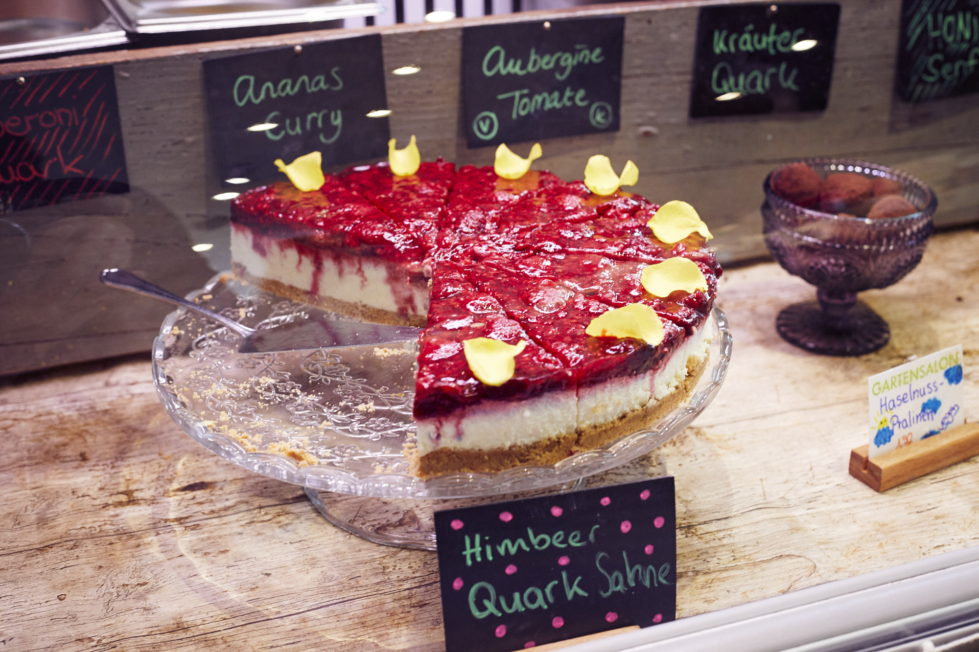 Himbeer-Quark-Kuchen auf einem Glasständer, dem ein Stück fehlt, mit gelben Blütenblättern obendrauf, ist der Hingucker in der Backwarenauslage – perfekt zum Feiern eines Junggesellenabschieds.