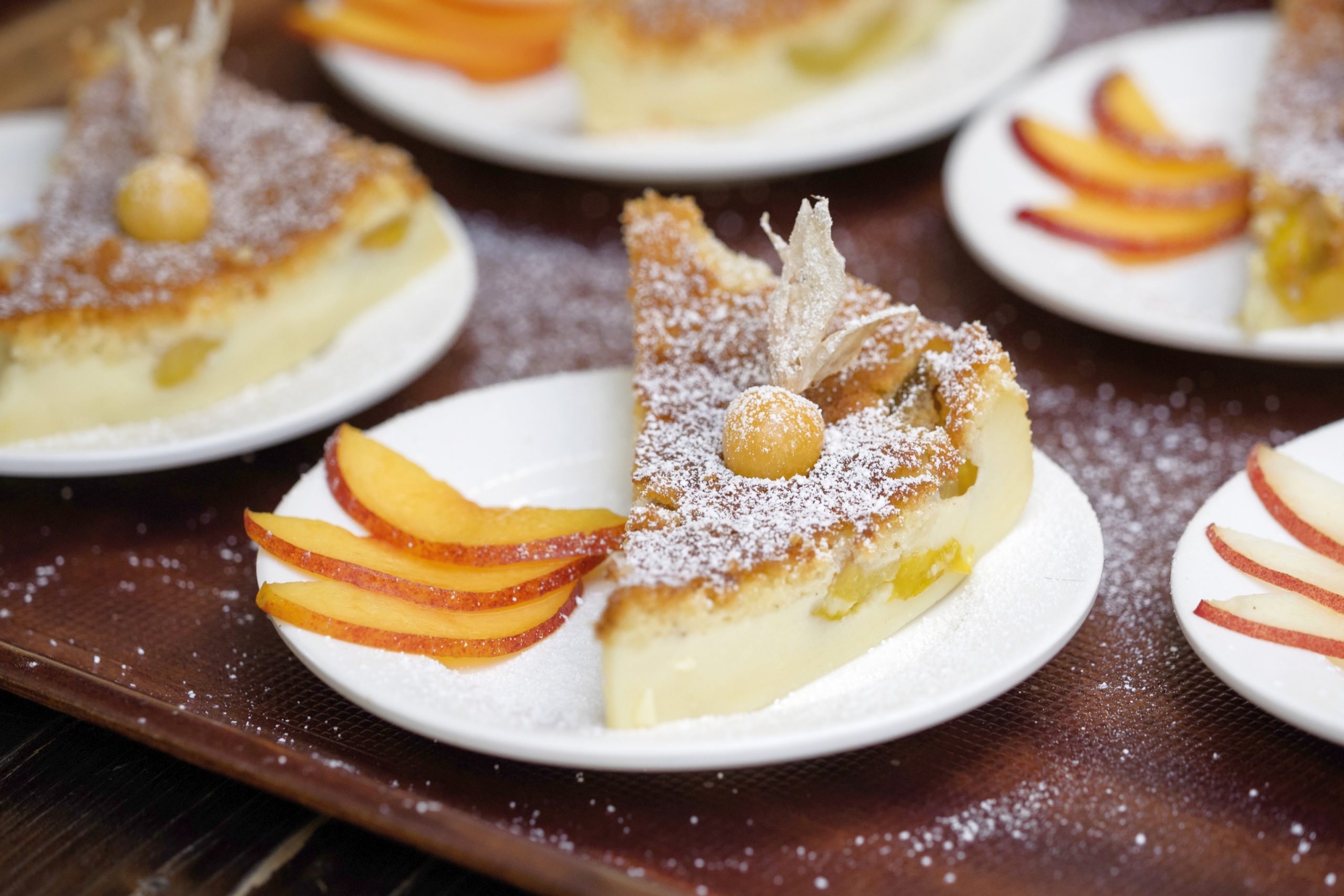 Mit Früchten belegte und mit Puderzucker überzogene Kuchenstücke, begleitet von Pfirsichscheiben auf weißen Tellern, sind ein köstlicher Leckerbissen, der sich perfekt für die Feier zum Junggesellenabschied eignet.