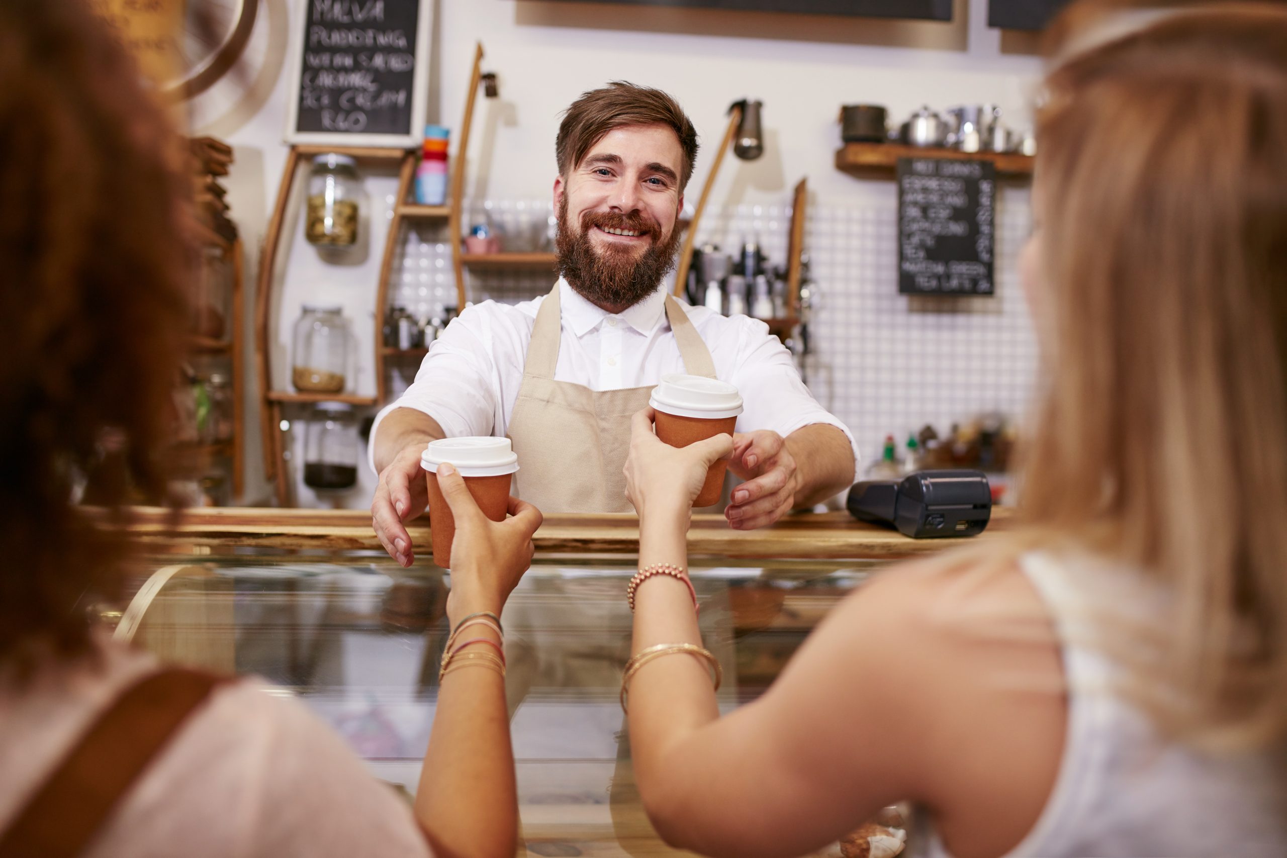 An der belebten Café-Theke überreicht ein bärtiger Barista, vielleicht gerade von einem vergnügten Junggesellenabschied zurück, mit einem freundlichen Lächeln den Kunden zwei dampfende Tassen Kaffee.