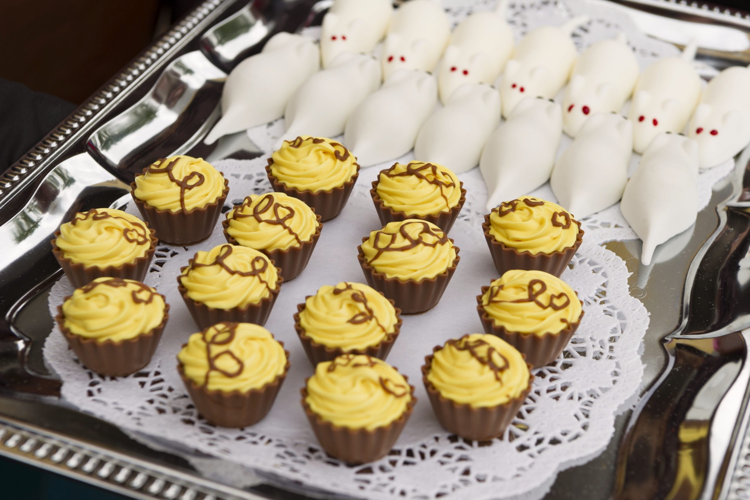 Ein Tablett mit Cupcakes mit gelbem Zuckerguss und weißen Süßigkeiten in Mäuseform mit roten Augen ist elegant auf einem Spitzendeckchen arrangiert, perfekt für eine Junggesellenabschiedsfeier.