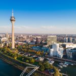 Luftaufnahme der Düsseldorfer Skyline mit dem Rheinturm und den Gehry-Bauten am Fluss unter einem klaren blauen Himmel.