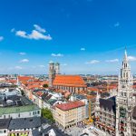 Luftaufnahme der Münchner Stadtlandschaft mit Marienplatz und Frauenkirche unter einem klaren blauen Himmel.