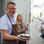 Mann hält einen Teller Brownies mit Schlagsahne auf einer Straße und lächelt, im Hintergrund sind zwei Personen zu sehen.