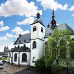 Historisches Kirchengebäude mit Türmen, umgeben von grünen Bäumen, unter einem blauen Himmel mit Wolken.