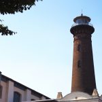 Hoher Leuchtturm aus Backstein vor einem klaren blauen Himmel, umgeben von Bäumen und einem Teil eines weißen Gebäudes.