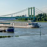 Ein Kreuzfahrtschiff fährt auf einem Fluss unter einer grünen Hängebrücke hindurch, im Hintergrund die Skyline einer Stadt.