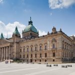 Historisches Gebäude mit großer Kuppel und Säulen vor blauem Himmel mit Wolken. Menschen gehen im Vordergrund.