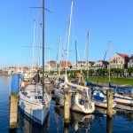 Yachten und ein Großsegler liegen in einem Yachthafen vor Anker, im Hintergrund sind Gebäude und ein klarer blauer Himmel zu sehen.