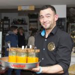 Lächelnder Mann hält ein Tablett mit Gläsern gelber Suppe in einem gemütlichen Café-Ambiente.