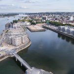 Luftaufnahme einer Uferpromenade mit Gebäuden, Brücken und einem Fluss unter einem teilweise bewölkten Himmel.