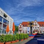 Ein Stadtplatz mit modernen und historischen Gebäuden, Pflanzkübeln, orangefarbenen Sonnenschirmen und einer geometrischen Skulptur unter einem blauen Himmel.