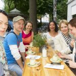 Eine Gruppe lächelnder Menschen sitzt in einem Straßencafé und genießt Getränke an einem Holztisch.