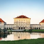 Ein großer Palast mit roten Dächern, die sich bei Sonnenuntergang in einem ruhigen Teich spiegeln.