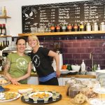 Zwei lächelnde Mitarbeiter hinter einer Café-Theke mit Gebäck und Menütafeln, in Regalen aufgereihte Weinflaschen.