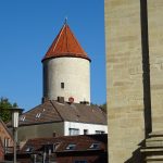 Ein runder Turm mit rotem Dach hinter einer Reihe von Gebäuden unter einem klaren blauen Himmel.