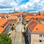 Luftaufnahme einer europäischen Stadt mit orangefarbenen Dächern, Menschen, die auf einer zentralen Straße spazieren, und einer Kirche im Hintergrund.