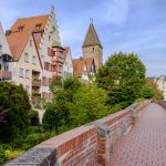 Ein Kopfsteinpflasterweg neben charmanten mittelalterlichen Häusern unter einem klaren Himmel, mit einem hohen Kirchturm im Hintergrund.