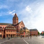 Panoramablick auf einen historischen europäischen Platz mit einer Kathedrale, farbenfrohen Gebäuden und einem wolkig blauen Himmel.