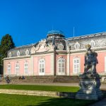 Eleganter rosa Palast mit verziertem Dach und Statue in einem sonnendurchfluteten Garten unter einem klaren blauen Himmel.