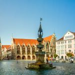 Stadtplatz mit einem Brunnen, historischen Gebäuden, einer Kirche und einem klaren blauen Himmel im Hintergrund.