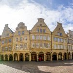 Ein sonniger Platz mit historischen Gebäuden, Bögen und einem hohen Kirchturm im Hintergrund.