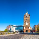 An einem sonnigen Tag mit klarem blauen Himmel steht inmitten moderner Gebäude ein historischer Uhrenturm.