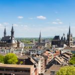 Panoramablick auf eine Stadtlandschaft mit historischen Kirchen, Dächern und Bäumen unter einem blauen Himmel mit vereinzelten Wolken.