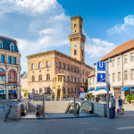 Ein sonniger Stadtplatz mit historischen Gebäuden, einem zentralen Uhrturm, Café-Sitzplätzen und einem U-Bahn-Eingang.