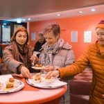 Menschen in warmer Kleidung genießen Essen von Tellern in einem hell erleuchteten Raum mit roten Wänden.