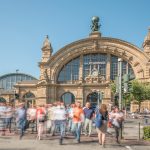 Verschwommene Menschenmenge vor einem historischen Bahnhof mit einer großen Uhr und einer Kuppel, unter einem klaren blauen Himmel.