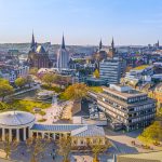 Luftaufnahme der Aachener Stadtlandschaft mit ihren historischen Gebäuden, üppigen Bäumen und einem belebten Platz an einem klaren, sonnigen Tag.