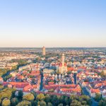 Luftaufnahme der charmanten Stadt Augsburg mit ihren ikonischen Gebäuden mit roten Dächern, einer hoch aufragenden Kirche und üppigem Grün unter einem klaren Himmel.