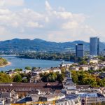 Eine Luftaufnahme von Bonn zeigt eine Stadtlandschaft mit einem gewundenen Fluss, modernen Gebäuden und Hügeln in der Ferne unter einem teilweise bewölkten Himmel.