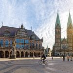 Eine Person fährt mit dem Fahrrad über einen sonnigen, historischen Platz mit reich verzierten Gebäuden und einer Kirche im Hintergrund.