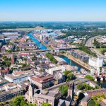 Luftaufnahme von Duisburg, einer Stadt, in der sich Flüsse anmutig durch Brücken schlängeln und eine Mischung aus modernen und historischen Gebäuden stolz unter einem klaren blauen Himmel aufragt.