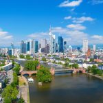 Die Luftaufnahme von Frankfurt zeigt eine Skyline mit Wolkenkratzern, einen ruhigen Fluss, der sich durch die Stadt schlängelt, und eine harmonische Mischung aus moderner und traditioneller Architektur unter einem klaren blauen Himmel.