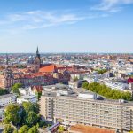 Die Luftaufnahme von Hannover zeigt eine Stadtlandschaft mit historischen Gebäuden, einer bezaubernden Kirche und üppigen Grünflächen unter einem klaren blauen Himmel.