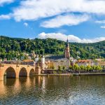 Erleben Sie einen atemberaubenden Panoramablick auf Heidelberg, Deutschland, wo die historische Alte Brücke anmutig den Neckar überspannt und ein majestätisches Schloss auf einem Hügel stolz unter dem weiten blauen Himmel thront.