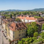 Die Luftaufnahme von Kassel zeigt eine charmante europäische Stadt mit Gebäuden mit roten Dächern, von Bäumen gesäumten Straßen und malerischen Hügeln im Hintergrund.